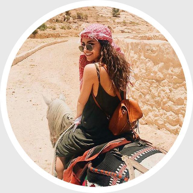 Person sitting outdoors, wearing a headscarf, with a bag next to them.