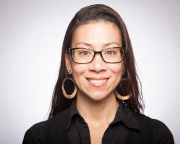 A person wearing glasses and earrings, smiling against a neutral background.