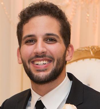 A person smiling, wearing a suit with a white boutonnière