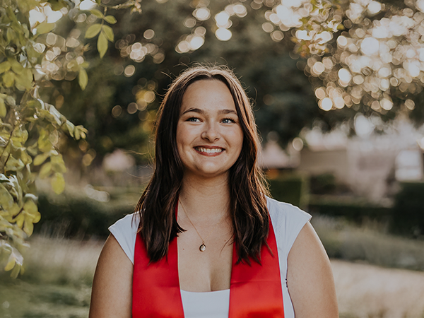 A young woman in a white shirt and red stole poses outside in the late evening.