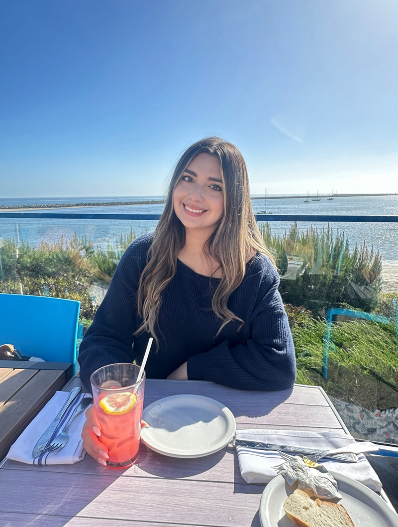 Alexis sits at an outdoor dining table with the water and sky in the background