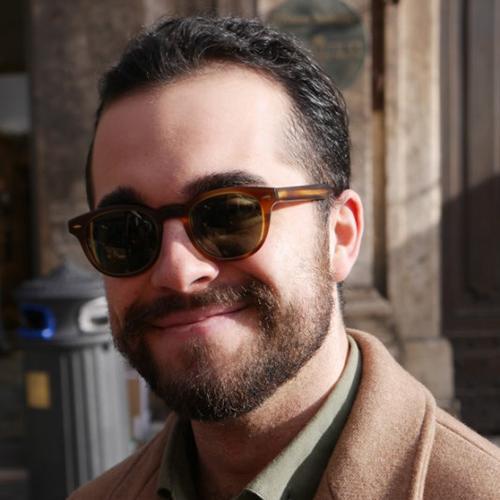 A person wearing sunglasses and a coat smiling outdoors.