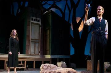 Two people acting on a stage with a dark, dramatic set. 