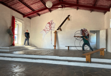 2 men stand on the left and 1 man stands on the left of a large room with cream walls, a wooden roof and grey tiled floor. Theres a bench on the right and modern art on the far wall.
