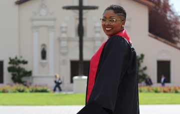 Person in graduation attire on a college campus, smiling at the camera.