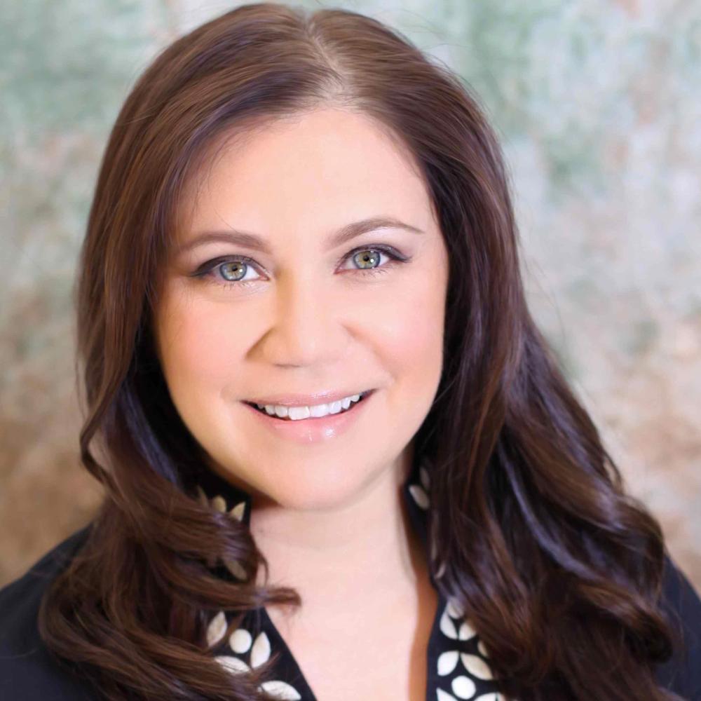 A smiling person with long hair wearing a dark blazer and patterned top.