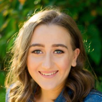 A smiling person wearing a denim jacket with greenery in the background.