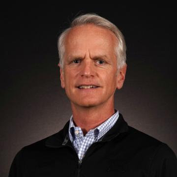 A person in a jacket and collared shirt with a dark background. image link to story