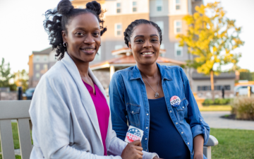 Two Black women sitting on a park bench. One is wearing a Vote pin and the other is holding an I voted sticker. Pocstock/Canva for Education