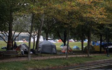 Multiple tents line the edges of a tree-lined city park near a Whole Foods grocery store. Image by Monica via Adobe Stock. 