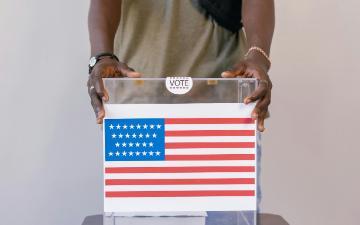 Person holding a ballot box with image of U.S. flag on the side. By Mikhail Nilov via Pexels. 