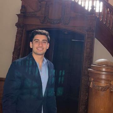 A person in a suit stands near a wooden staircase and window.