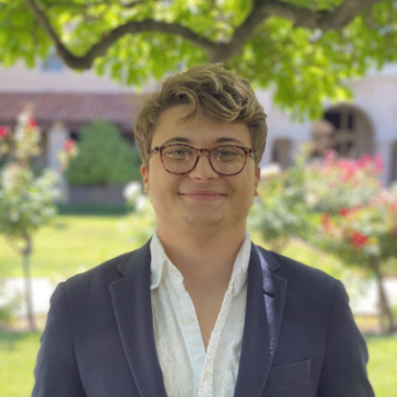A person wearing a suit standing outdoors with trees and bushes in the background.