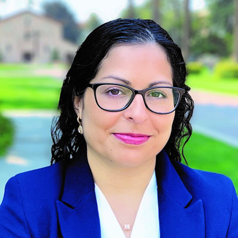 A person wearing glasses and a blazer, with houses and trees in the background.