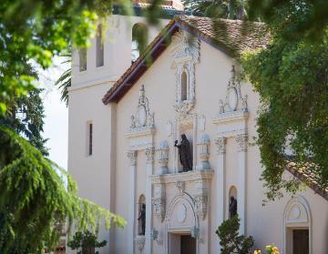 The image shows the front view of Mission Santa Clara surrounded by greenery. image link to story