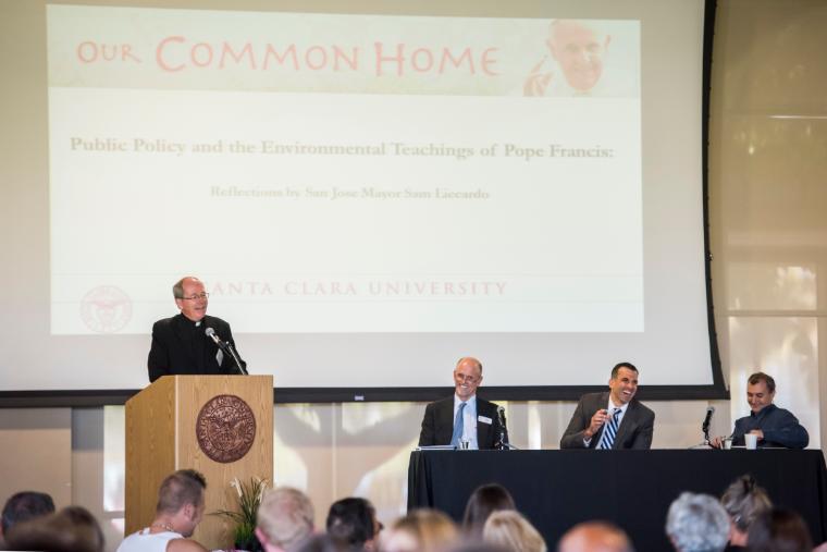 A speaker at a podium with three panelists in front of an audience.