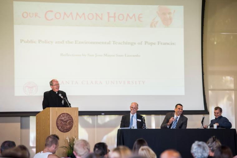 A speaker at a podium with three panelists in front of an audience.
