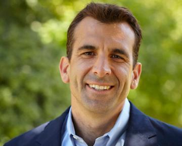 A man smiling in an outdoor setting with greenery in the background.
