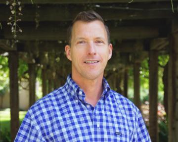 A person in a checked shirt standing outside under a wooden structure.