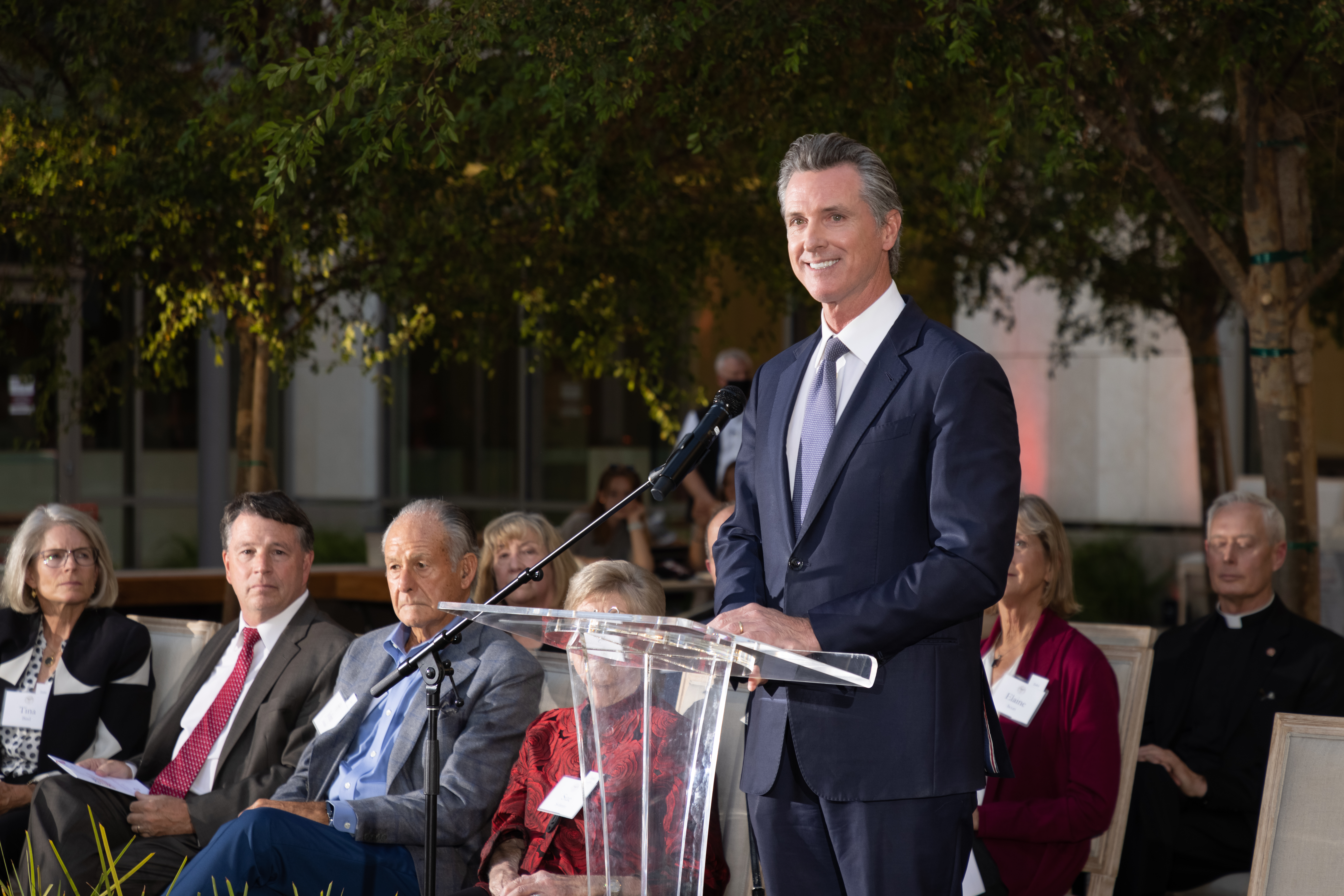 Gavin Newsom speaking at an outdoor dedication event with seated attendees.