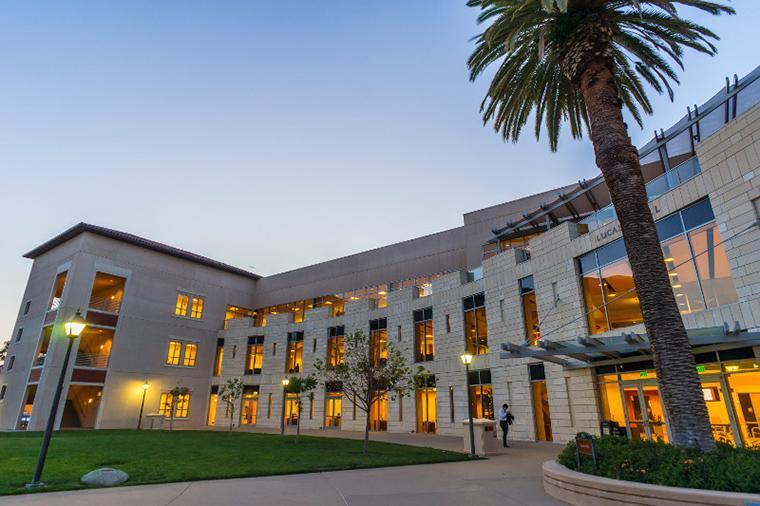 A large building labeled Lucas Hall with windows illuminated at dusk and a tall palm tree.