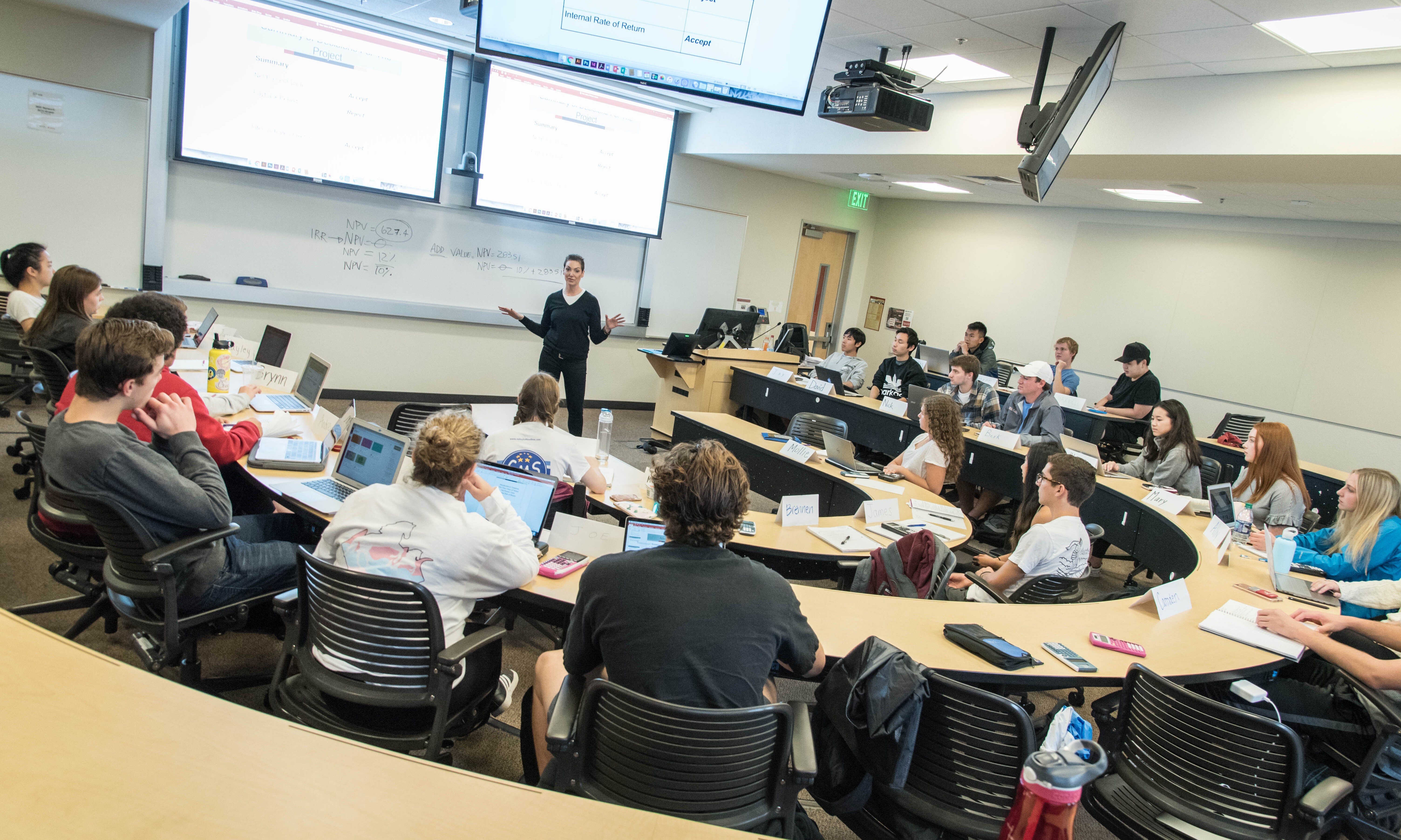 Professor lecturing to a full classroom of students with multiple screens displaying information.