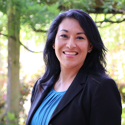 A woman in a black blazer outside with trees in the background.