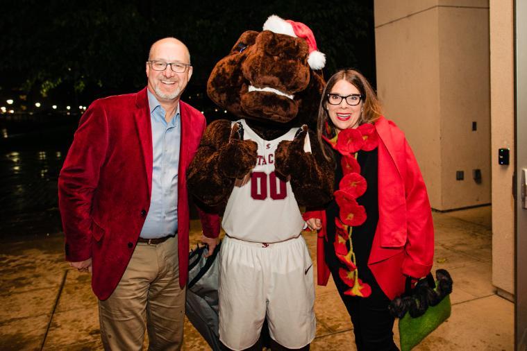 Two people pose with a festive mascot at an event.