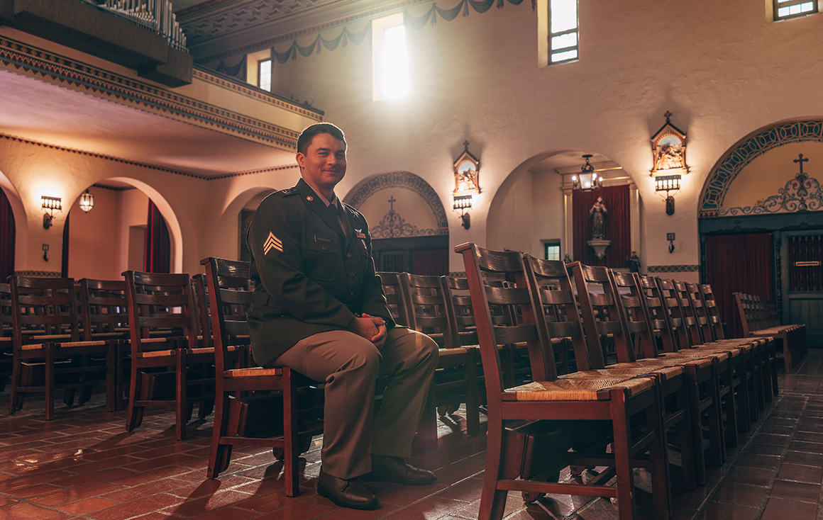 A young man in a military uniform sits in a wooden chair inside a historic church, the rising sun lighting him up from a window.