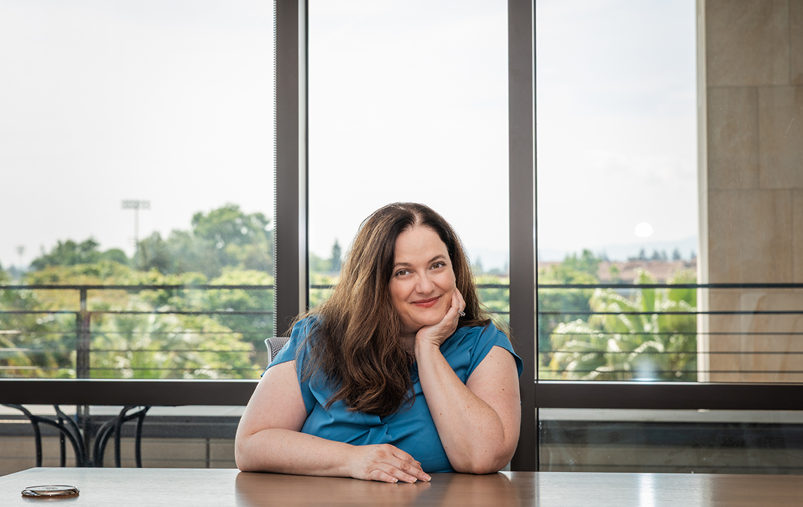 A woman in a blue blouse sits at a conference room table, one arm resting casually on the tabletop while she leans and rests her head on the palm of her propped arm. A view of the sky is visible through the room's dramatic, large windows.