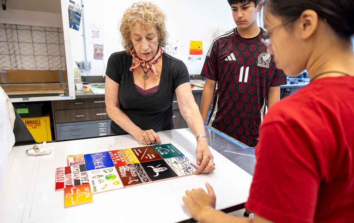 An older woman lays out a collection of linocuts atop a press, with two young people watching her.