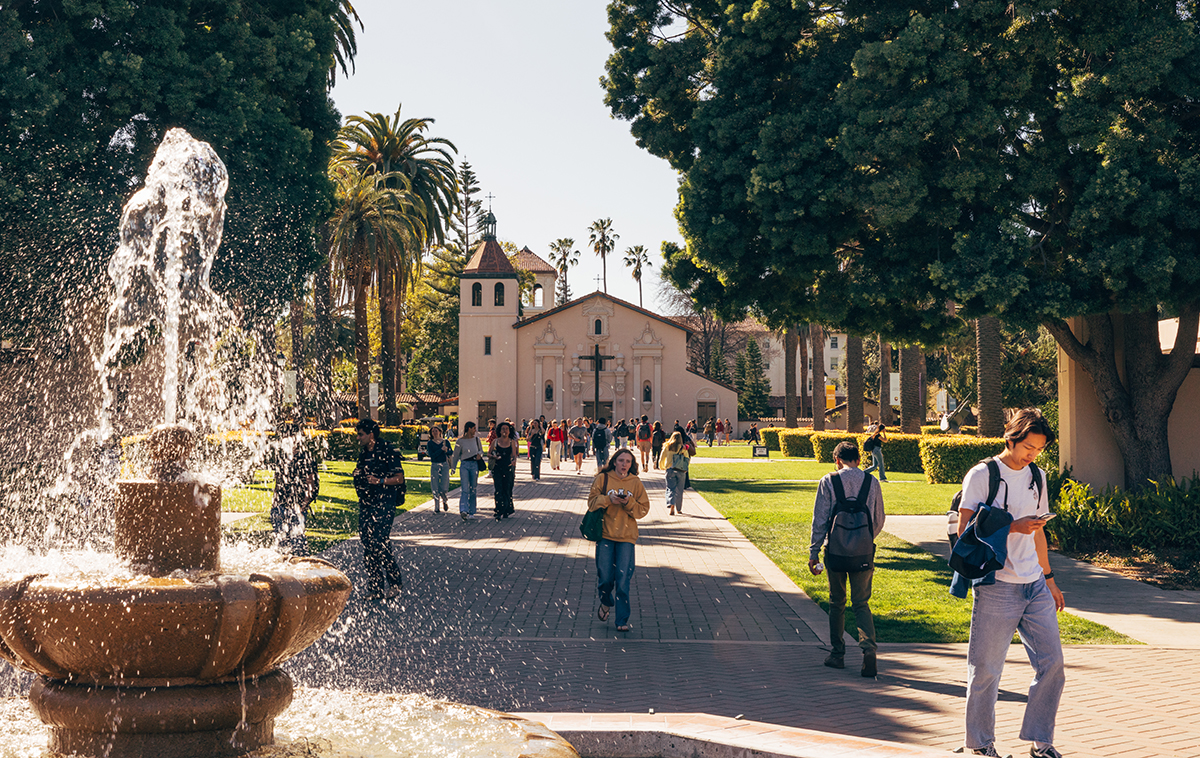 Students walking away from Mission Church on Palm Drive with water coming from fountain, illuminated by the sun.