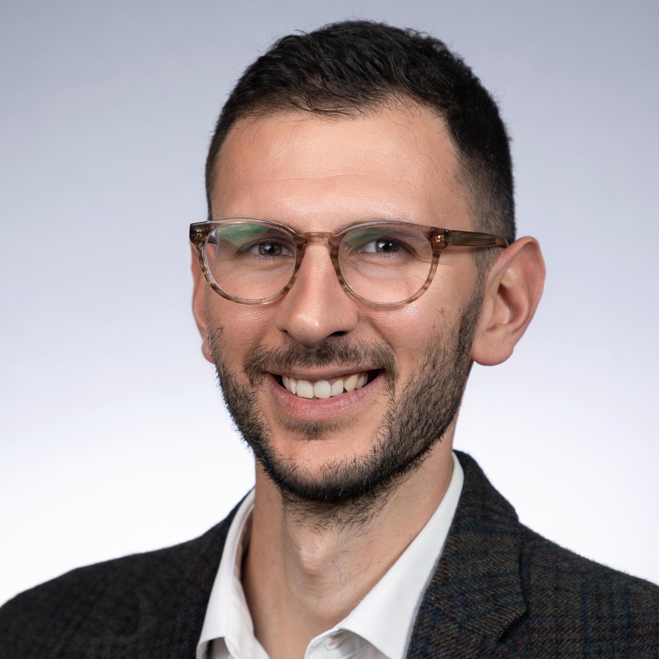 A man wearing glasses in a suit, smiling against a gray background.