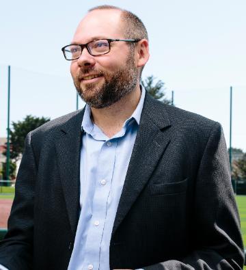 A person in a suit standing outdoors, holding a camera.