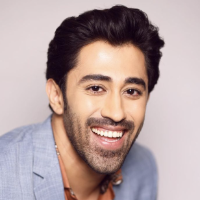 Smiling person in a light blue suit with a brown shirt.