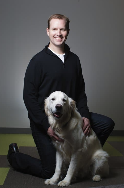 A man kneeling next to a golden retriever, both looking forward.