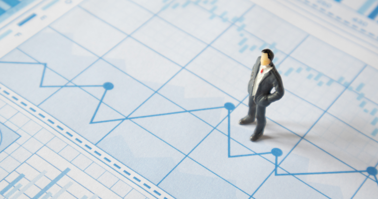 A miniature man in a gray suit stands on a light blue financial chart with various data lines and bars. image link to article