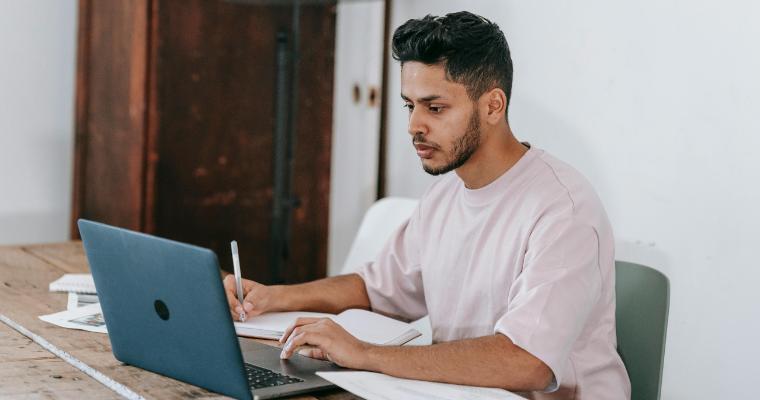 Student working on laptop while taking notes by hand