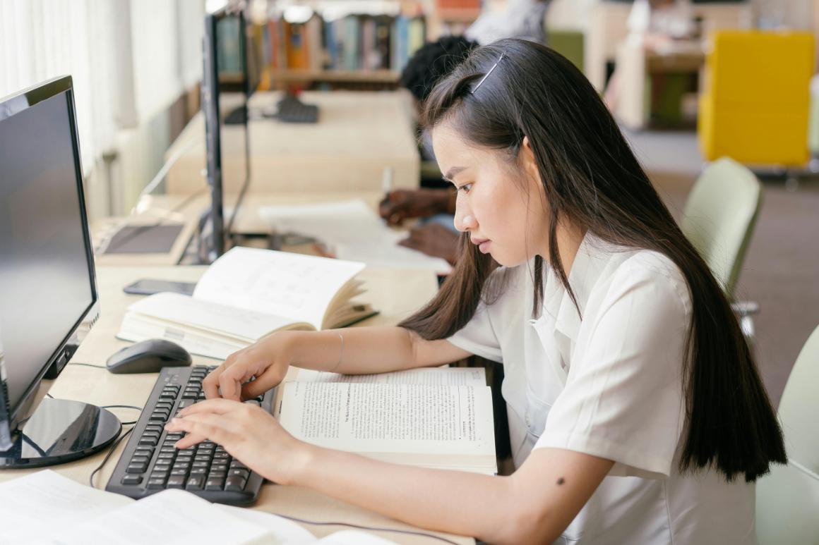 Student reading book while typing on desktop computer