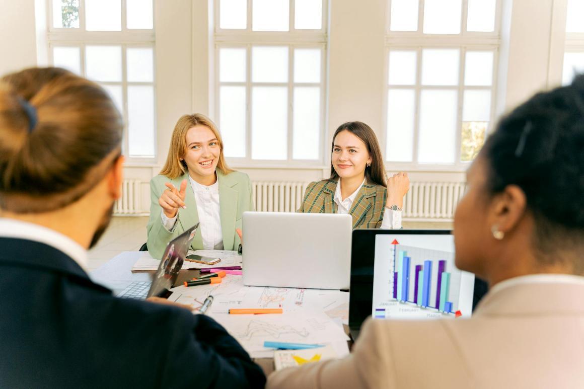 Two women are seated at a conference table, smiling and actively engaged in discussion with other team members who are partially visible in the foreground