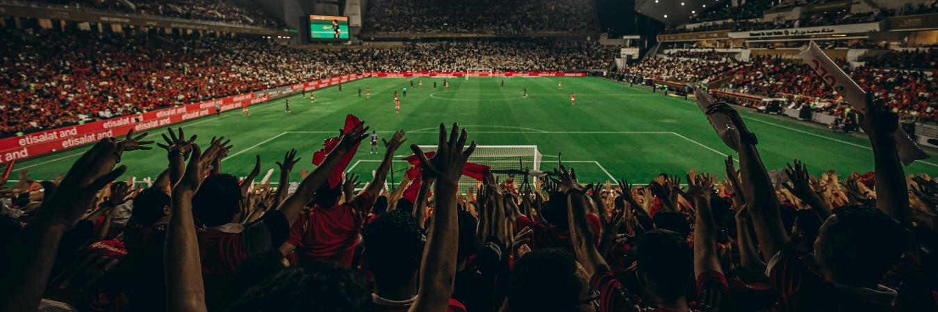 A vibrant football stadium at night, with fans in red jerseys cheering. The pitch is brightly lit, brimming with excitement and energy.