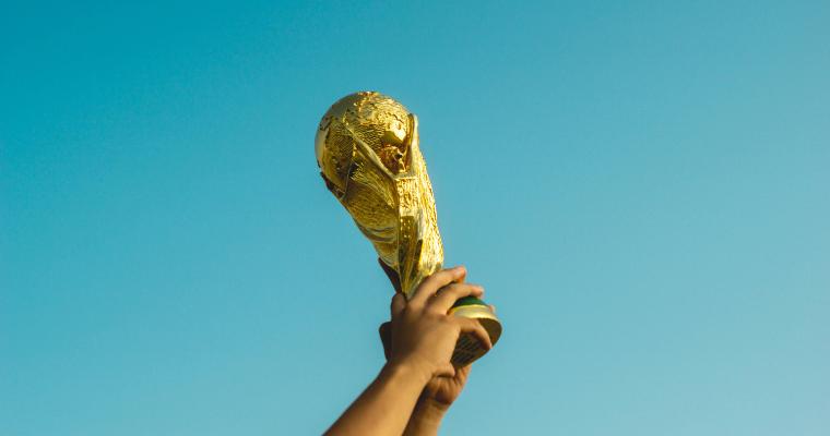 A golden trophy, resembling the FIFA World Cup, is held up against a clear blue sky. image link to article