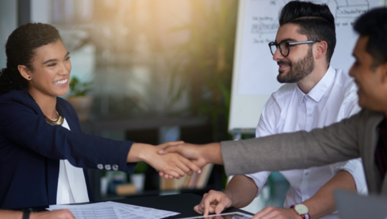 People in business attire around a table with two shaking hands.