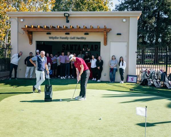 Two men take a swing on the putting green in front of a crowd at the entrance of a building with a sign that says Wipfler Family Golf Facility