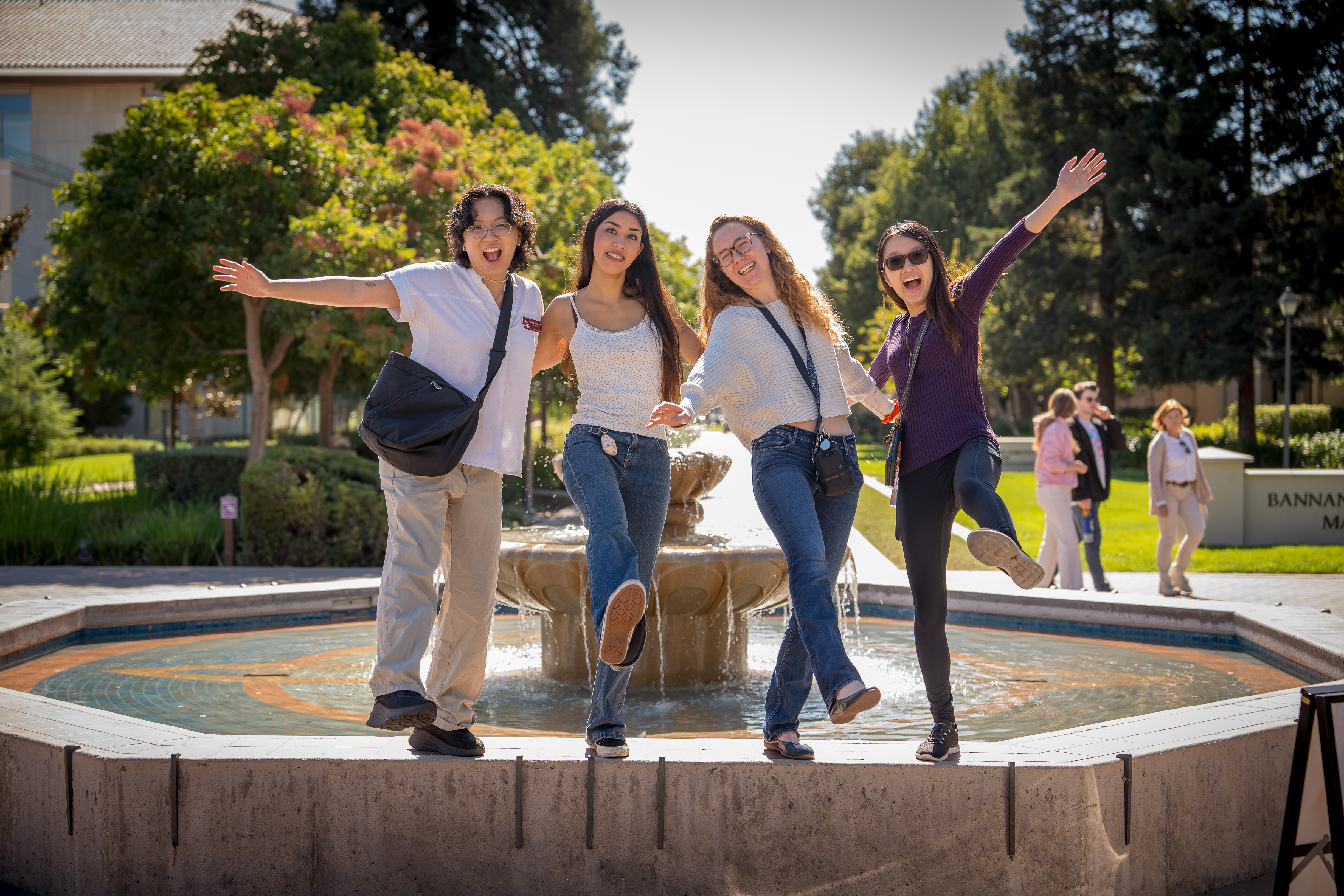 Four student tour guides stand on the edge of a fountain with their arms in the air and one foot kicked out.