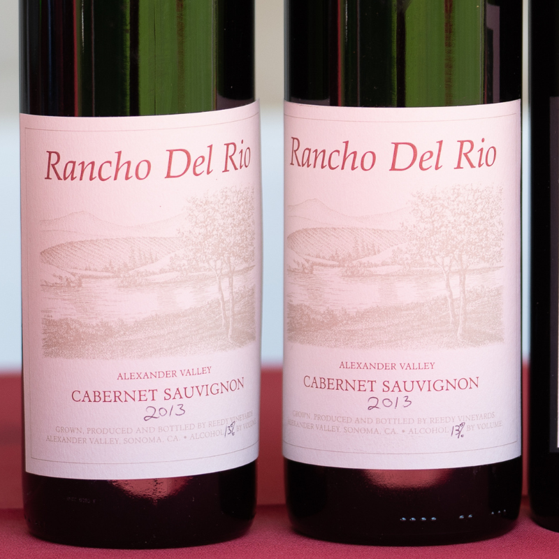 A close-up of labels on two wine bottles. The top reads Rancho del Rio above a drawing of two trees overlooking a lake in a valley. Vineyards line the slopes of the mountains. Beneath are the words  Alexander Valley Cabernet Sauvignon 2013 