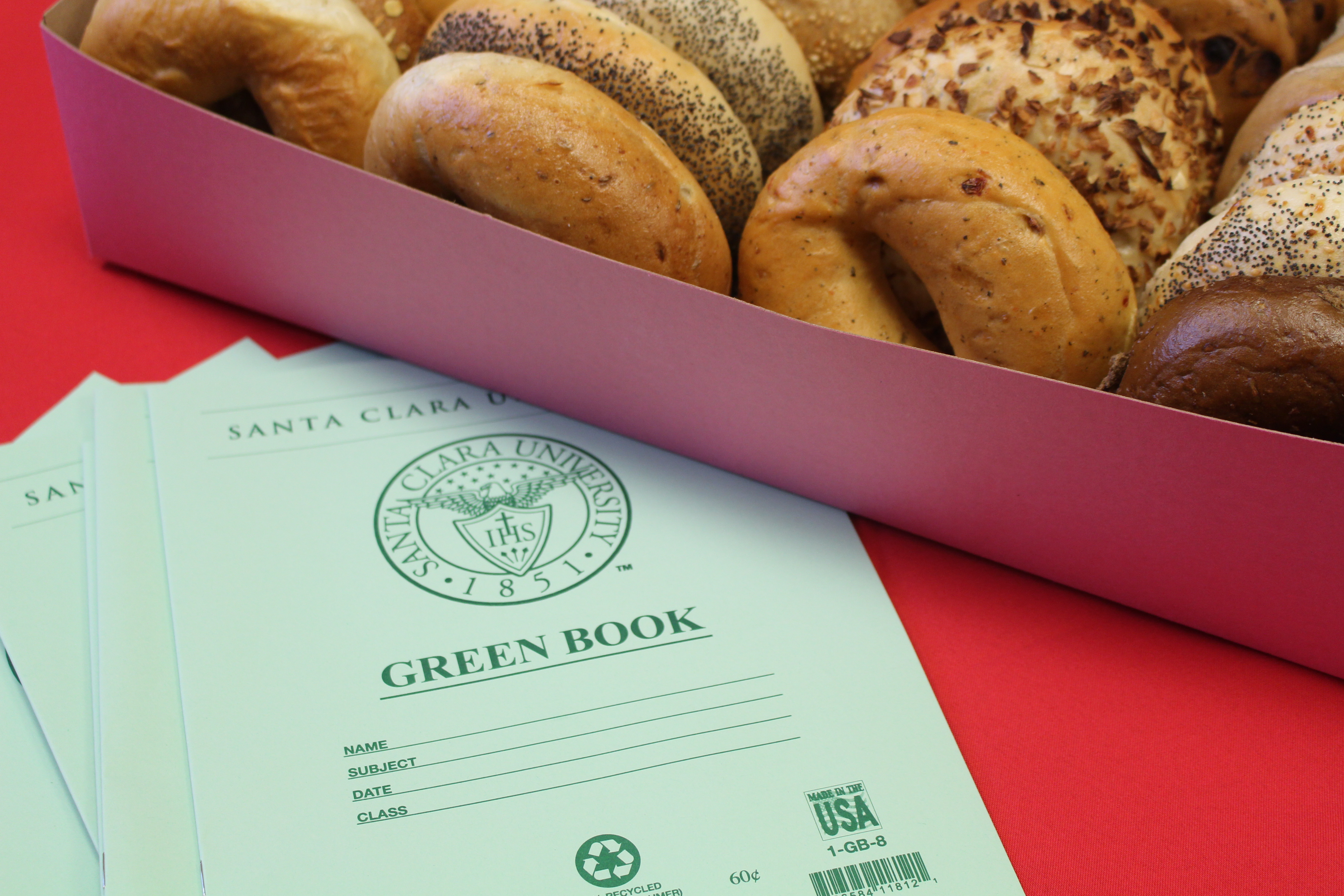 Box of assorted bagels next to green notebooks on a red surface.
