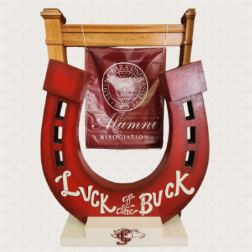 A large, red wooden horseshoe on a white base. White cursive text on the horseshoe reads Luck of the Buck. In the background is a red banner with the Santa Clara University seal and the words Alumni Association.