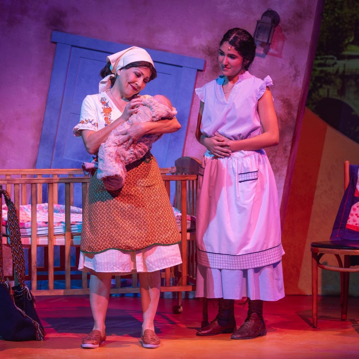 Two women in costume stand on a stage. The elder of the two is holding a swaddled baby, and both regard the child fondly.