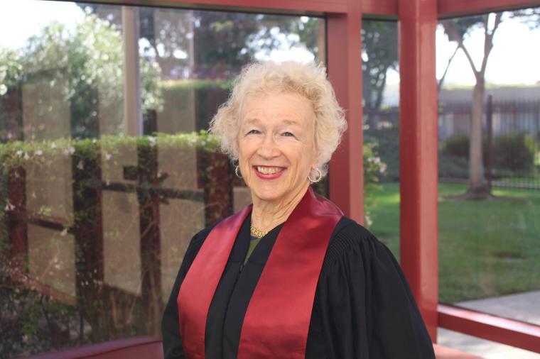 A person wearing a black robe with a red stole, outdoor background.